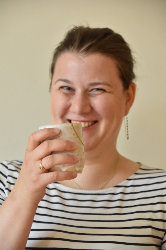 Frederike lächelt in die Kamera und hält einen Teebecher hoch.
