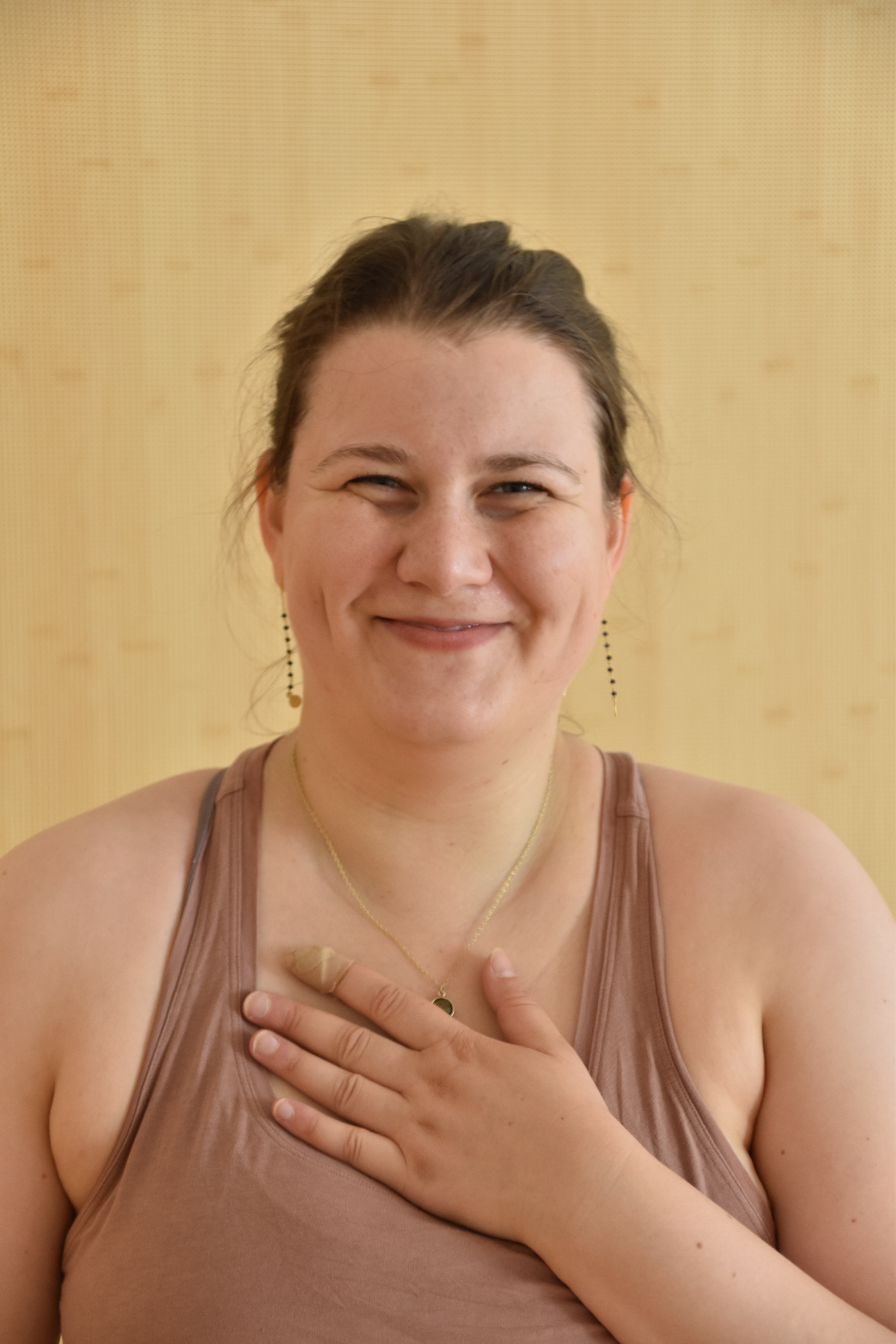 Frederike steht vor einer hellen Holzwand und hat ihre lange Hand auf ihr Herz gelegt