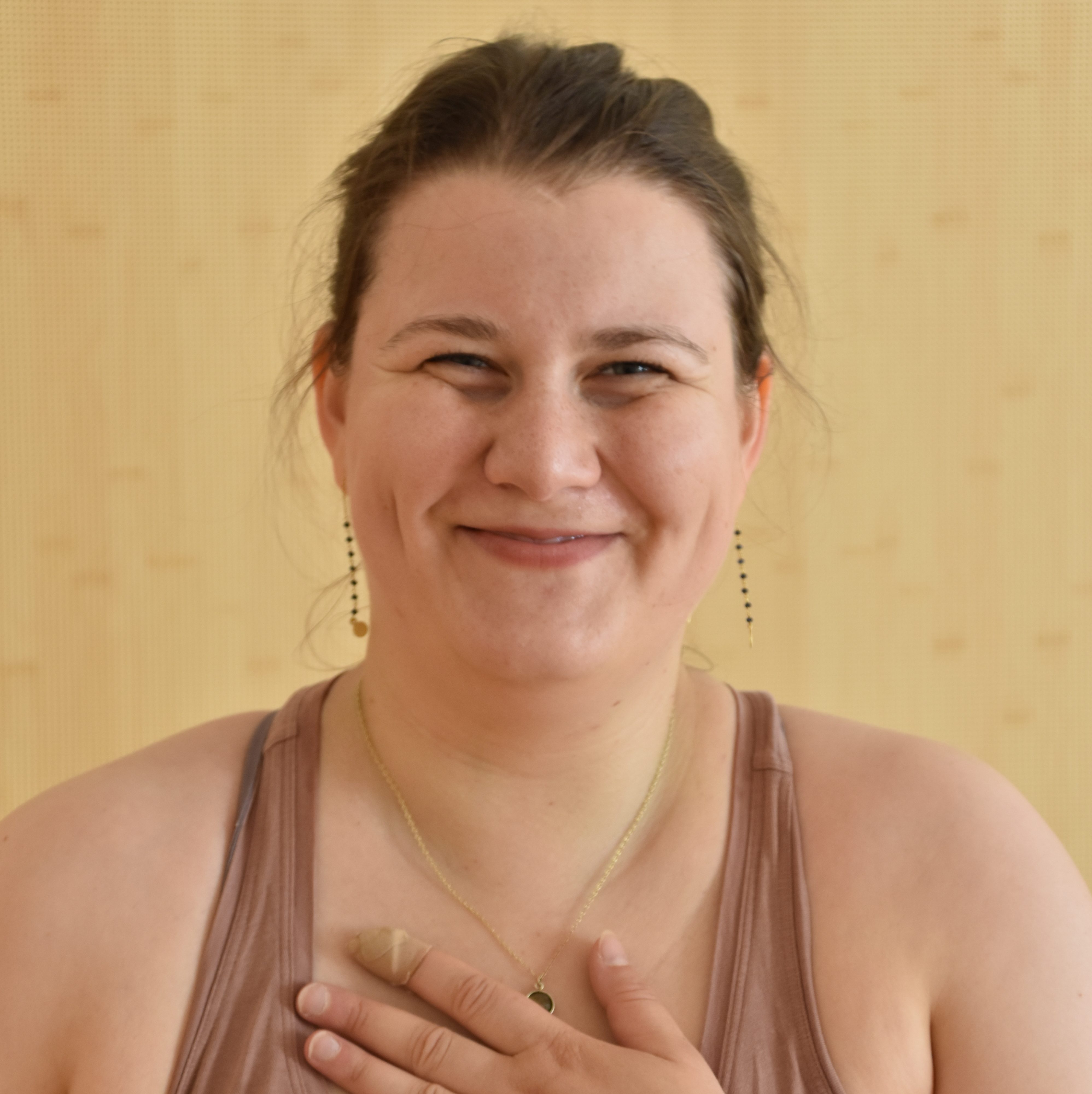 Frederike steht vor einer hellen Holzwand und hat ihre lange Hand auf ihr Herz gelegt